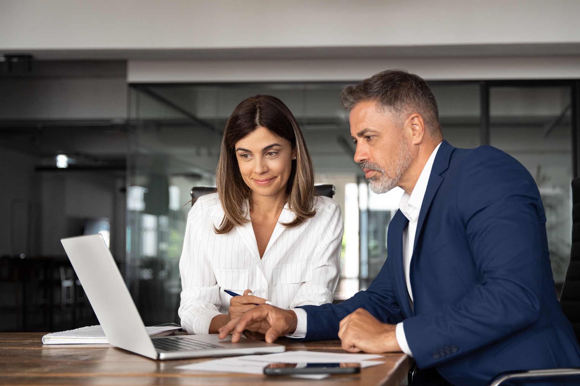 Team of diverse partners sitting at table mature Latin business man and European business woman discussing project on laptop in office. Two colleagues of professional business people working, close up
